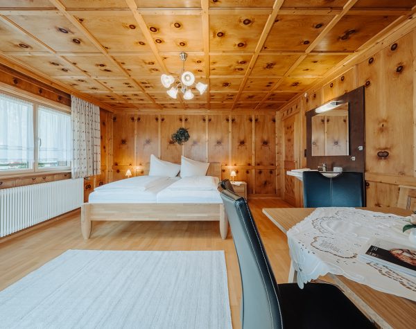 rustikales Schlafzimmer mit Holztafeln an Wand und Decke, Holzboden, helles Doppelbett aus Holz, Eckbank, Waschbecken und große Fensterfront