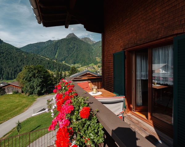 Blick von rustikalem Balkon mit Geranien auf Bergpanorama und Balkontür umgeben von Hauswand mit Holzschindeln