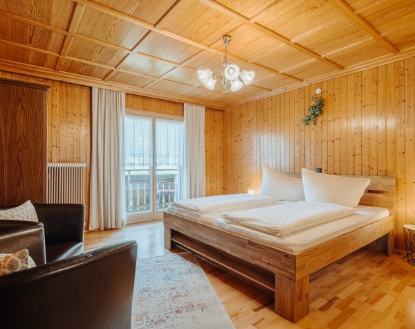 rustikales Schlafzimmer mit Holztafeln an Wand und Decke, Holzboden, Doppelbett aus Holz, Balkontür, Holzschrank und zwei Sessel am Fußende des Bettes