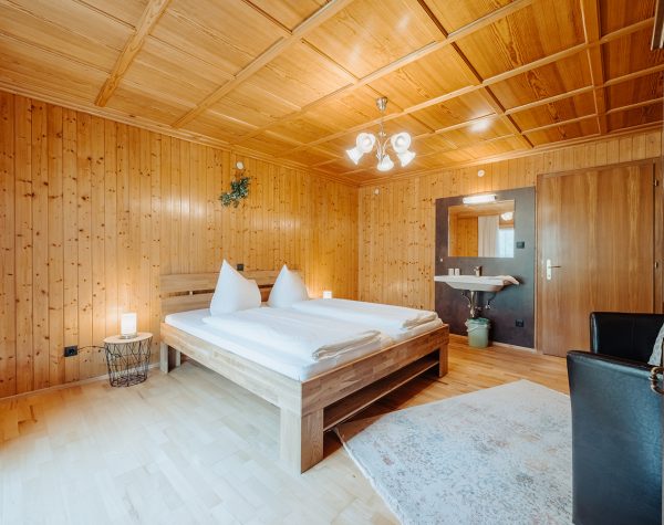 rustikales Schlafzimmer mit Holztafeln an Wand und Decke, Holzboden, Blick von Balkontür auf Doppelbett aus Holz, Zimmertür rechts des Waschbeckens und Sessel