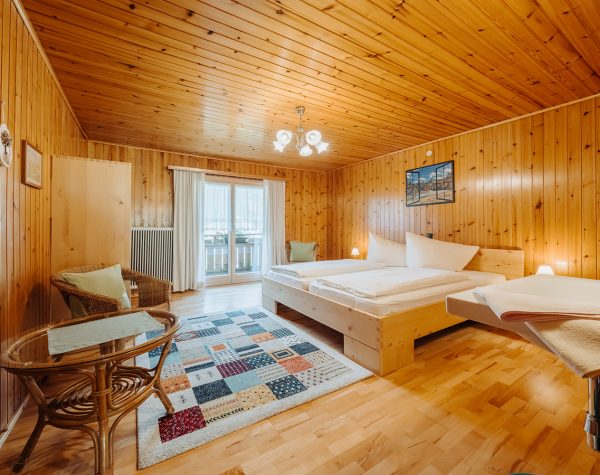 rustikales Schlafzimmer mit Holztafeln an Wand und Decke, Holzboden, helles Doppelbett aus Holz, Balkontür, kleinem Tisch und Stuhl und Waschbecken