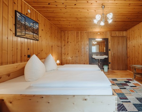 rustikales Schlafzimmer mit Holztafeln an Wand und Decke, Holzboden, helles Doppelbett aus Holz, Zimmertür, kleinem Tisch und Stuhl und Waschbecken
