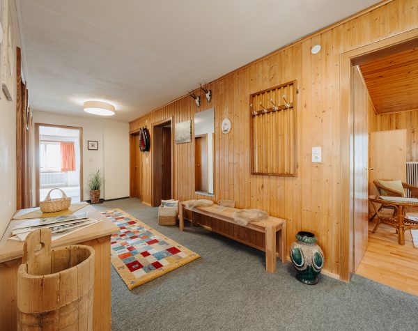 rustikaler dennoch heller Flur mit teils weißen Wänden und Holzvertäfelung, weiße Decke, grauer Teppichboden, Kommode, Holzbank und Blick ins Schlafzimmer und Küche