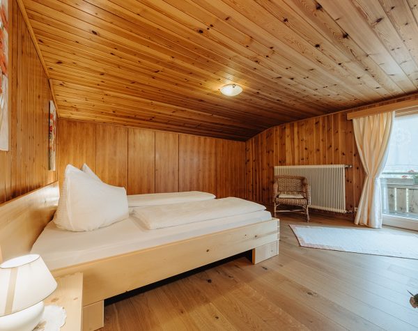 rustikales Schlafzimmer mit Holzvertäfelungen und Einbauschrank, helles Doppelbett mit Blick auf Balkontür, Nachttisch und Stuhl aus Korbgeflecht