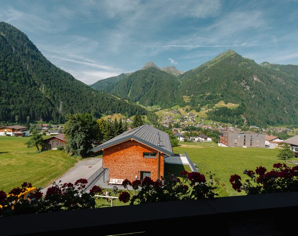 Ausblick vom mit Geranien gesäumten Balkon auf die sonnige Bergkulisse