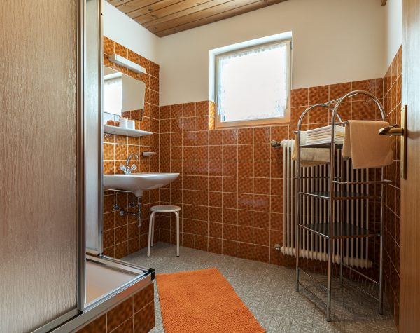 altes traditionelles Bad mit orange-braunen Wandfließen und grauem Boden, ausgestattet mit Dusche und Waschbecken und Heizkörper