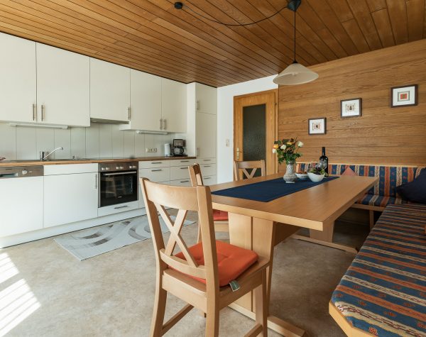 Küche mit dunkler Holzvertäfelung an Wand und Decke, hellem Boden, helle Küchenzeile und Eckbank mit Tisch und 3 Stühlen