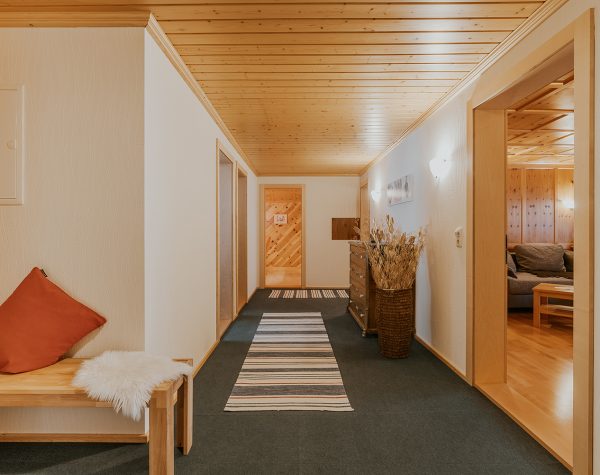 großer Flur mit weißen Wänden, grauem Teppichboden, Kommode und Blick geradeaus in die Küche und rechts vorn in das Wohnzimmer und vorne links eine Holzbank