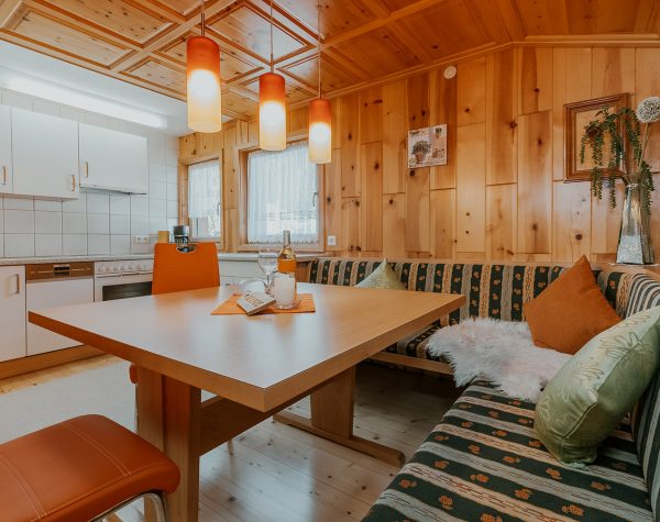 Wohnküche mit rustikaler Holzwand und Decke, Holzboden, Eckbank mit Tisch und 2 Stühlen und weiße Küche mit Fenstern neben der Küchenzeile