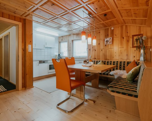 Wohnküche mit rustikaler Holzwand und Decke, Holzboden, Eckbank mit Tisch und 2 Stühlen und weiße Küche mit Fenstern neben der Küchenzeile