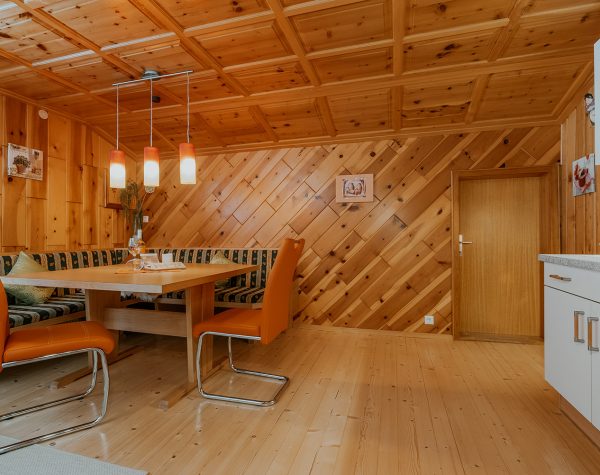 Wohnküche mit rustikaler Holzwand und Decke, Holzboden, Eckbank mit Tisch und 2 Stühlen und weiße Küche