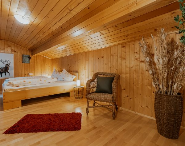 rustikales Schlafzimmer mit Holzvertäfelung an Decke und Wand, Holzoboden und Holzdoppelbett unter einer Dachschräge mit Nachttisch aus modernem Metallgestell und Stuhl aus Korbgeflecht