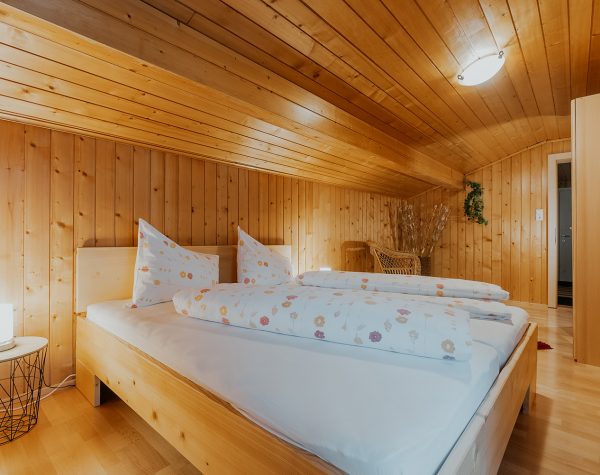 rustikales Schlafzimmer mit Holzvertäfelung an Decke und Wand, Holzoboden und Holzdoppelbett unter einer Dachschräge mit Nachttisch aus modernem Metallgestell