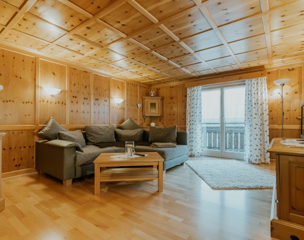 rustikales Wohnzimmer mit Holzvertäfelung an Wand und Decke von Wohnung 2, mit Schnitzereien verziertem Holzschrank und Kommode mit grauem modernen Sofa neben der Balkontür