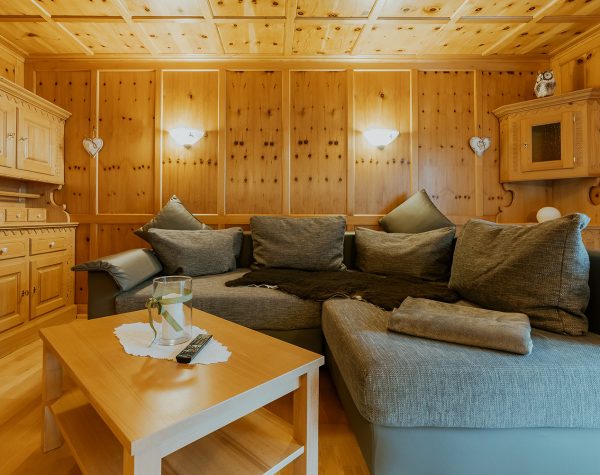 Wohnzimmer Wohnung 2 mit rustikaler Holzvertäfelung, mit Schnitzereien verzierter Holzschrank und grauem modernen Sofa im Hintergrund beleuchtet mit 2 Wandleuchten